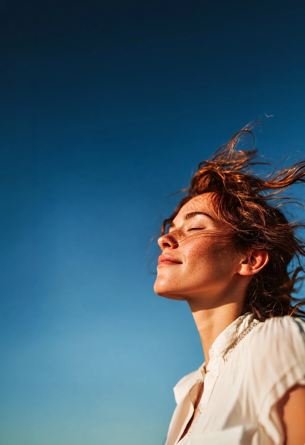 A person with eyes closed, hair moving in the wind against a clear blue sky